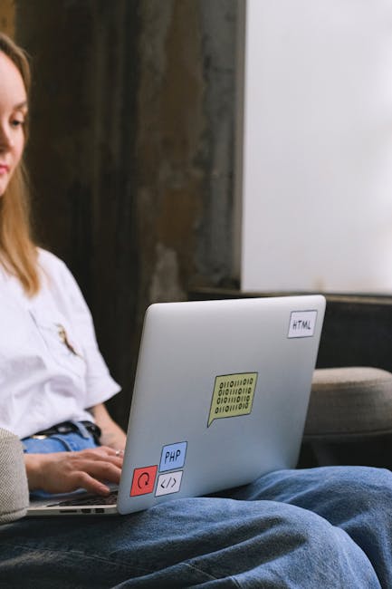 A person with long hair, dressed in a white top, is sitting outdoors on a bench or similar surface in a garden setting. They are using a silver laptop, which features several colorful stickers, includ