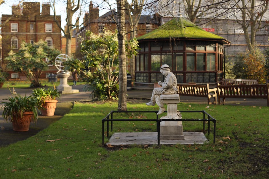 A person with long hair, dressed in a white top, is sitting outdoors on a bench or similar surface in a garden setting. They are using a silver laptop, which features several colorful stickers, includ