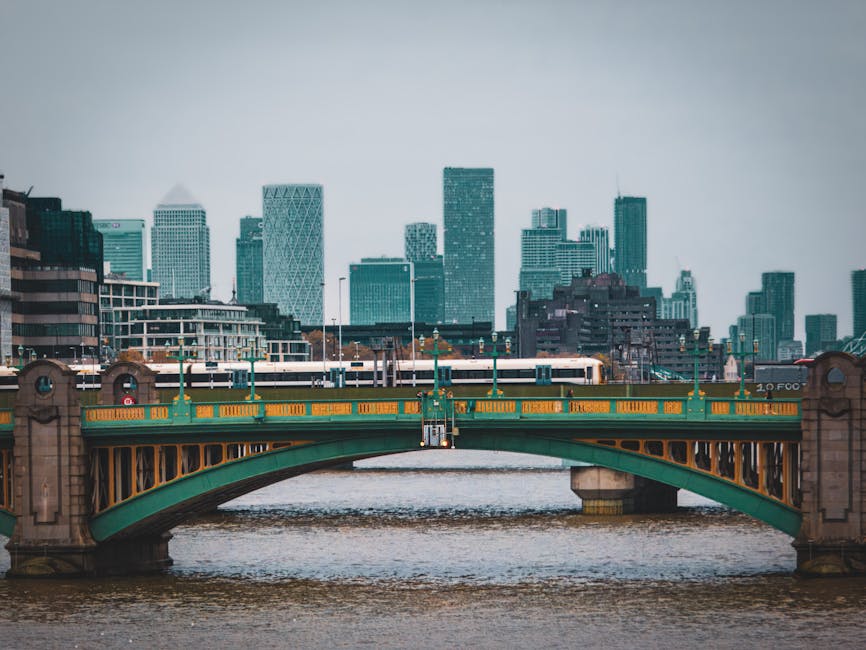 A view of a cityscape with tall modern skyscrapers and office buildings in the background, featuring a green and yellow Victorian-style bridge spanning a river in the foreground. A white train is cros