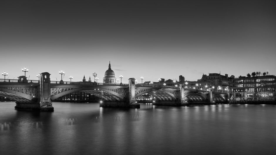 A view of a cityscape with tall modern skyscrapers and office buildings in the background, featuring a green and yellow Victorian-style bridge spanning a river in the foreground. A white train is cros