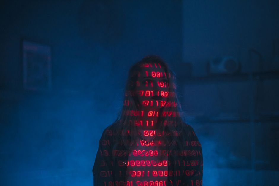 A person standing in a dimly lit room with a dark background, their face and body obscured by a hooded sweatshirt. The sweatshirt displays red digital code or binary numbers projected across the fabri